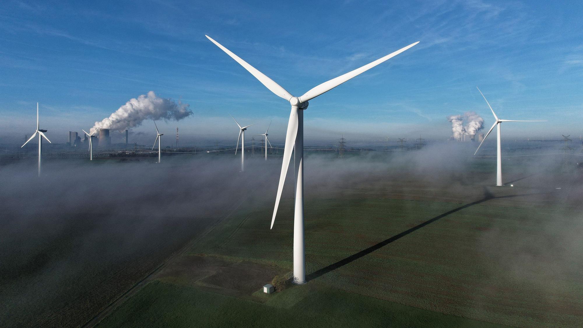 Windräder stehen auf einer Anhöhe nahe Berghaim, ein Kraftwerk ist in der Ferne zu sehen.