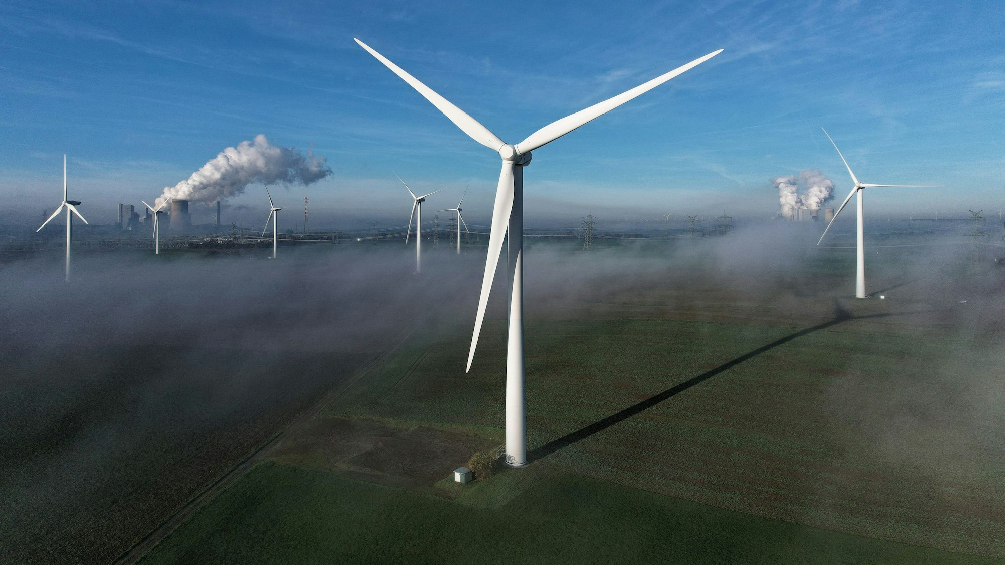 Auf dem Bild sind mehrere Windräder in Bergheim zu sehen, im Hintergrund auch zwei Kohlekraftwerke.