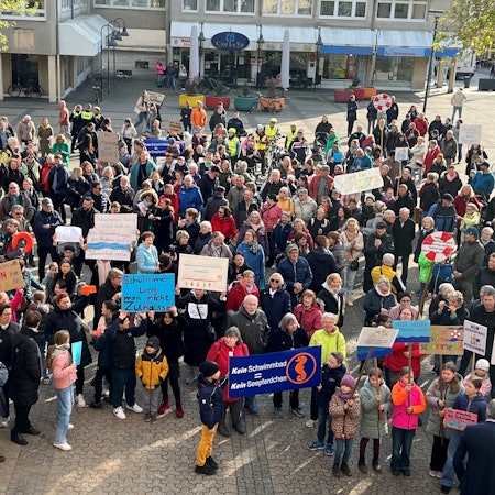 Auf dem Foto sind protestierende Menschen zu sehen. Sie halten Plakate gegen die Schließung des Schulschwimmbads hoch.