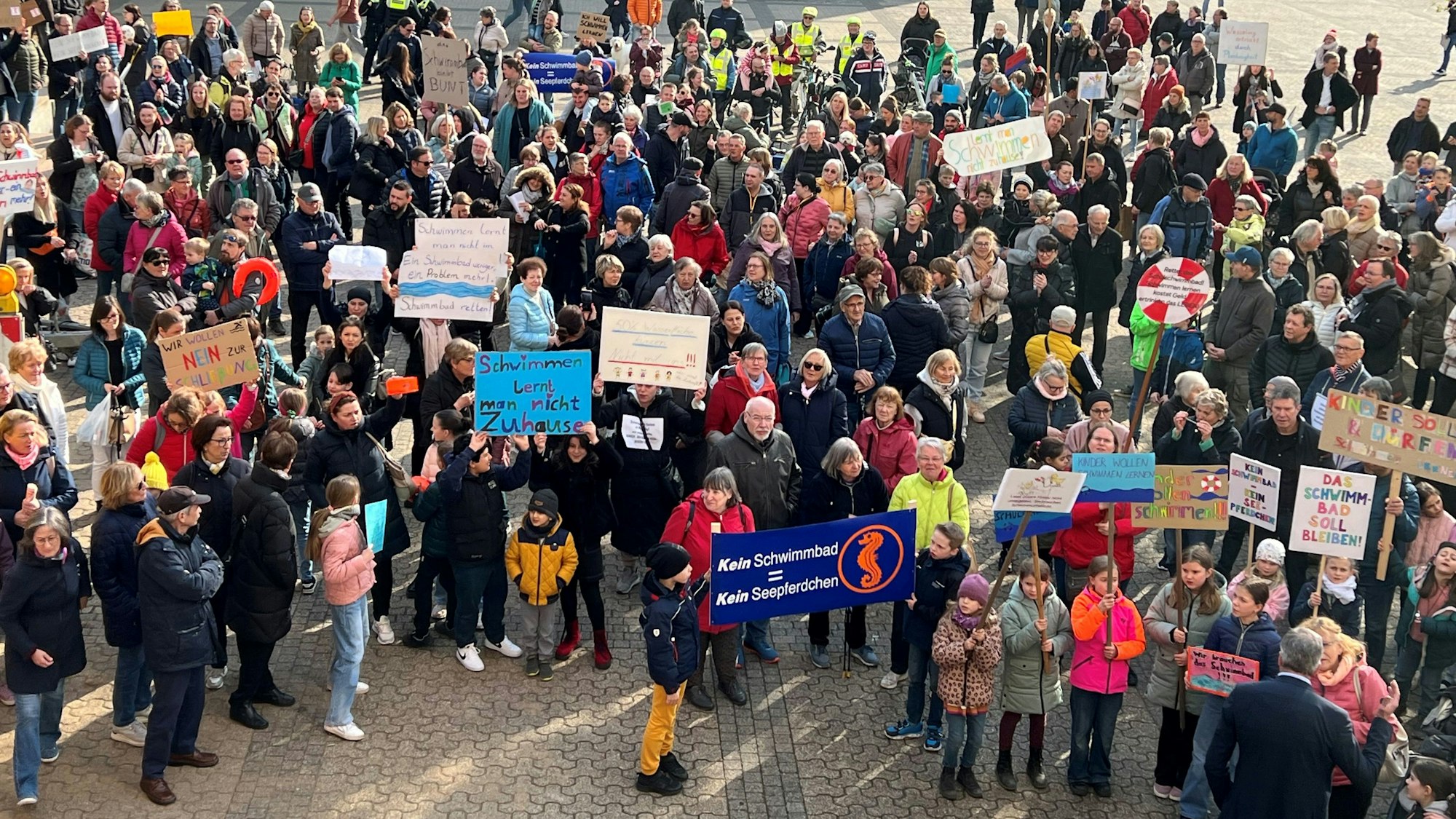 Auf dem Foto sind protestierende Menschen zu sehen. Sie halten Plakate gegen die Schließung des Schulschwimmbads hoch.