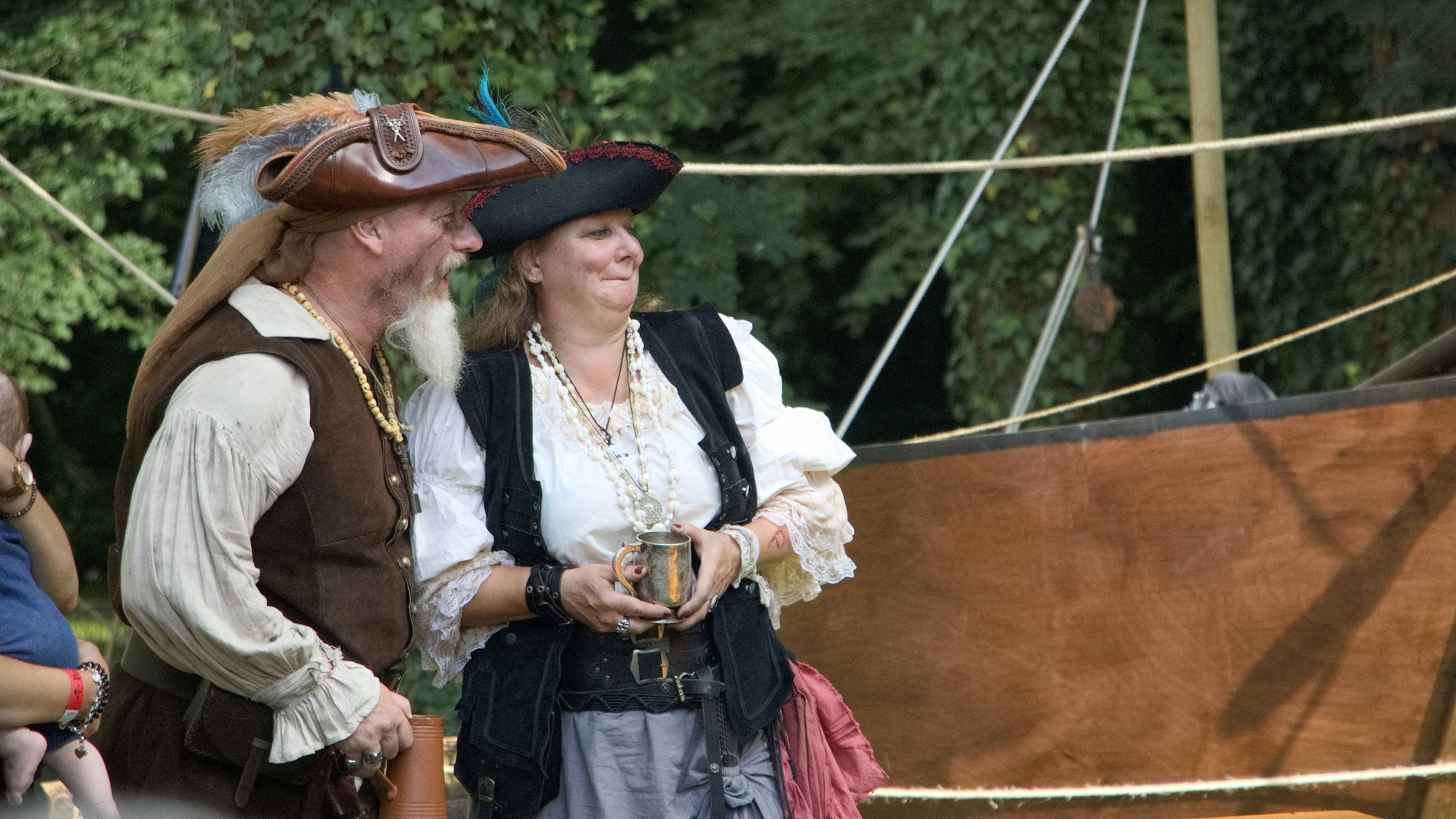 Ein Mann und eine Frau, selbst wie Piraten kostümiert, verfolgen vor einem Holzboot das Geschehen.