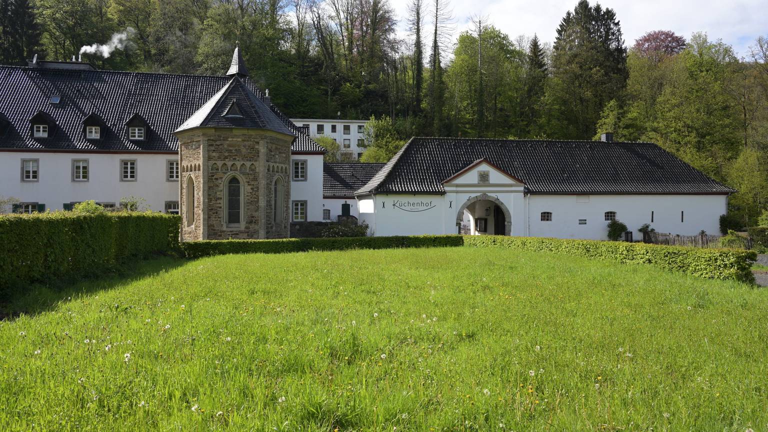 Eine Kapelle in einer Wiese, dahinter ein Hotel und ein historischer Küchenhof.