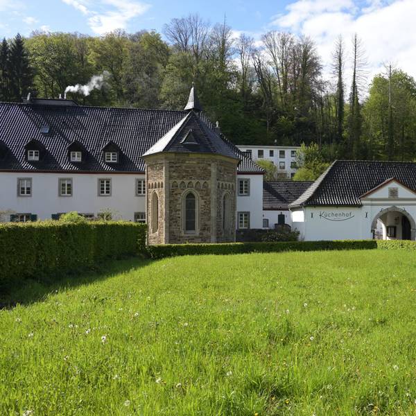 Eine Kapelle in einer Wiese, dahinter ein Hotel und ein historischer Küchenhof.