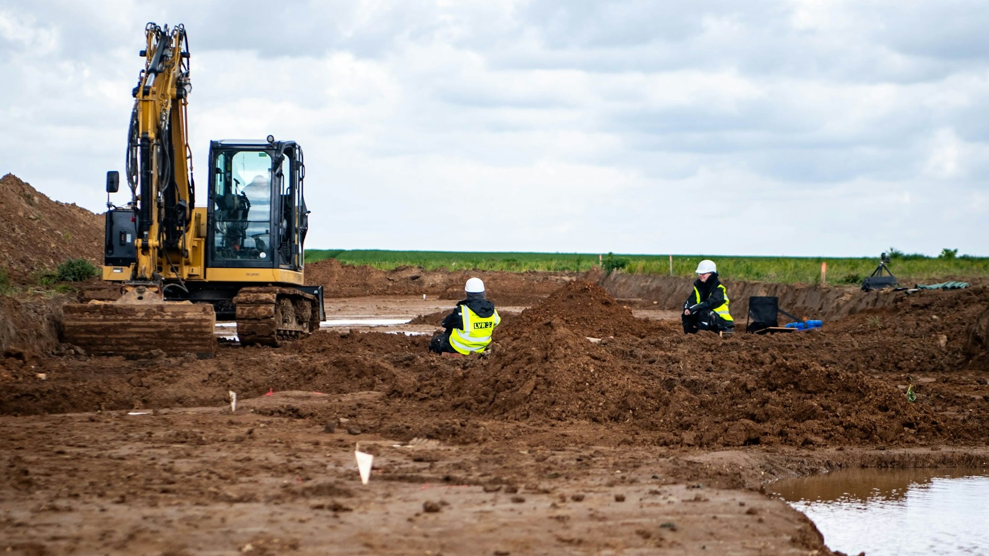 Das Bild zeigt zwei Mitarbeiter des LVR, die im Bereich der Grabungsfläche sitzen. Auch ein Bagger ist zu sehen.