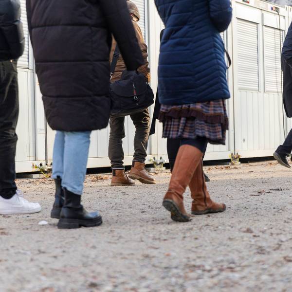 Die Beine von Menschen laufen über einen geschotterten Platz, im Hintergrund sind Wohncontainer für Geflüchtete zu sehen.