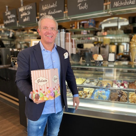 Andreas Siebrecht trägt einen Anzug. Vor der Eisauslage präsentiert er einen Pappkarton mit der Aufschrift „Gelato“.