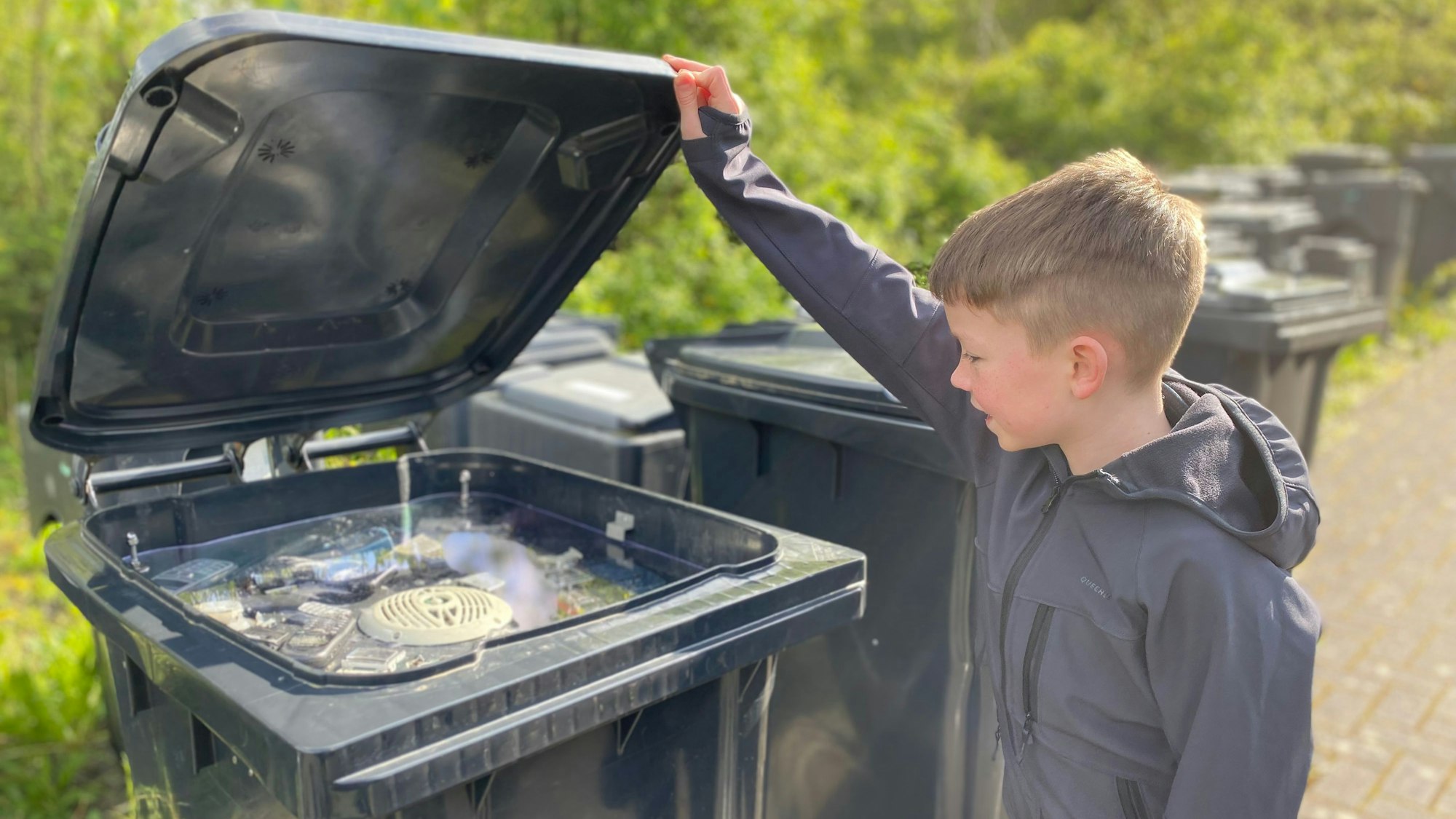 Leon öffnet eine Mülltonne. Darin befindet sich ein Schaukasten.