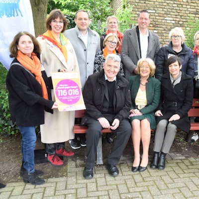 Zu sehen ist eine Gruppe von Menschen auf und rund um eine Bank.