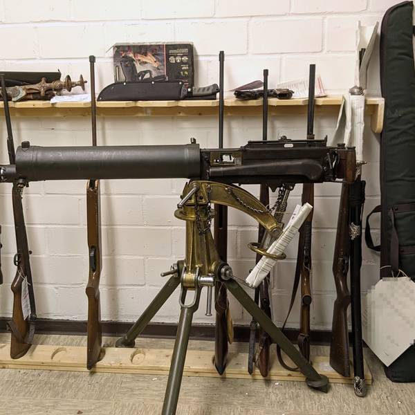 Ein altes Maschinengewehr ist auf einem Dreibein montiert. Dahinter lehnen diverse Waffen an der Wand.