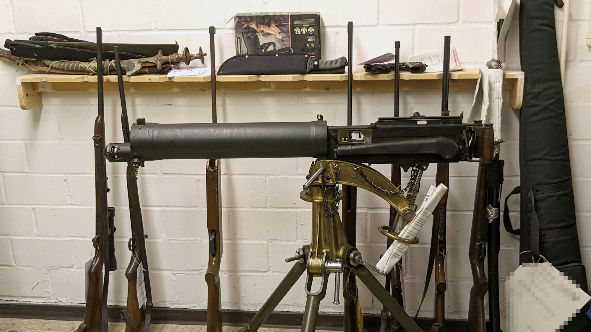Ein altes Maschinengewehr ist auf einem Dreibein montiert. Dahinter lehnen diverse Waffen an der Wand.