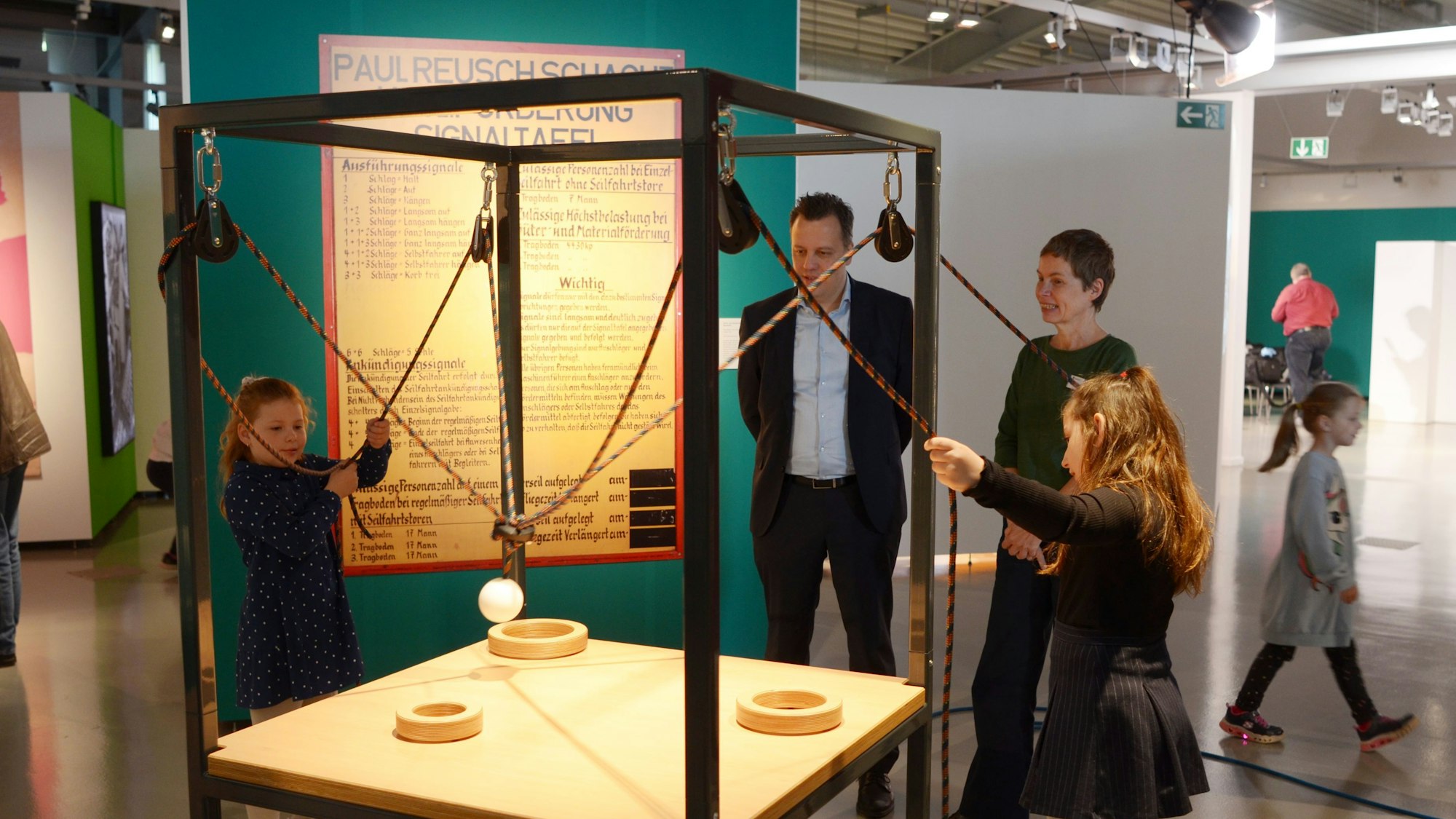 Zwei Mädchen steuern mit Seilzügen einen Ball. Er soll in einem der drei unterschiedlich großen Ringe landen, die auf einer Tischplatte platziert sind.