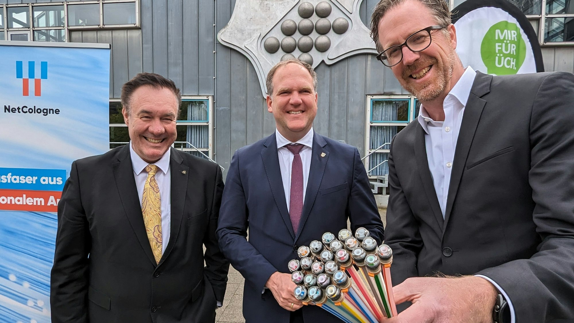Drei Männer im Anzug stehen vor dem Hürther Bürgerhaus, einer zeigt ein aufgefächertes Bündel an Glasfaserleitungen.