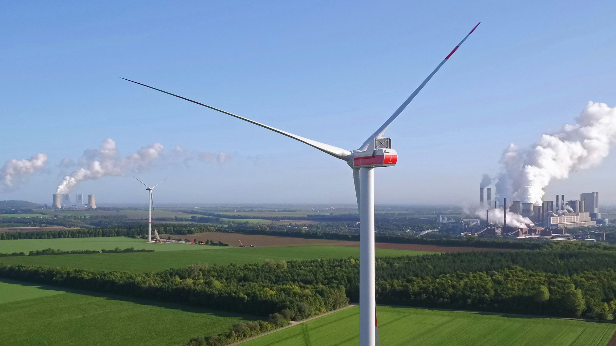 Zwei Windräder, im Hintergrund sind Kraftwerke zu sehen.