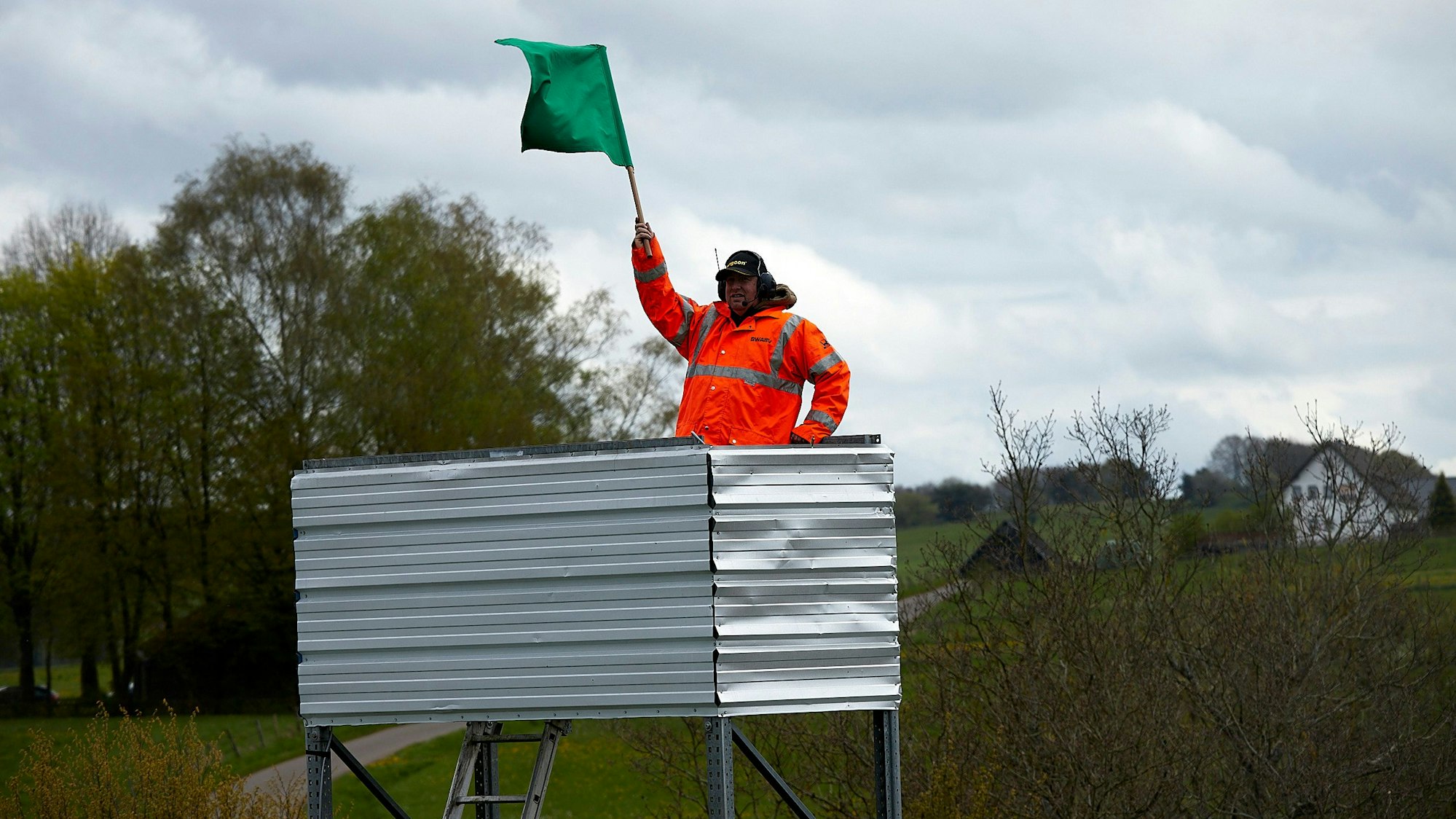 Ein Mann in einer signalfarbenen Jacke steht auf einem erhöhten, mit Aluminiumblenden verkleideten Podest und schwenkt eine grüne Fahne.