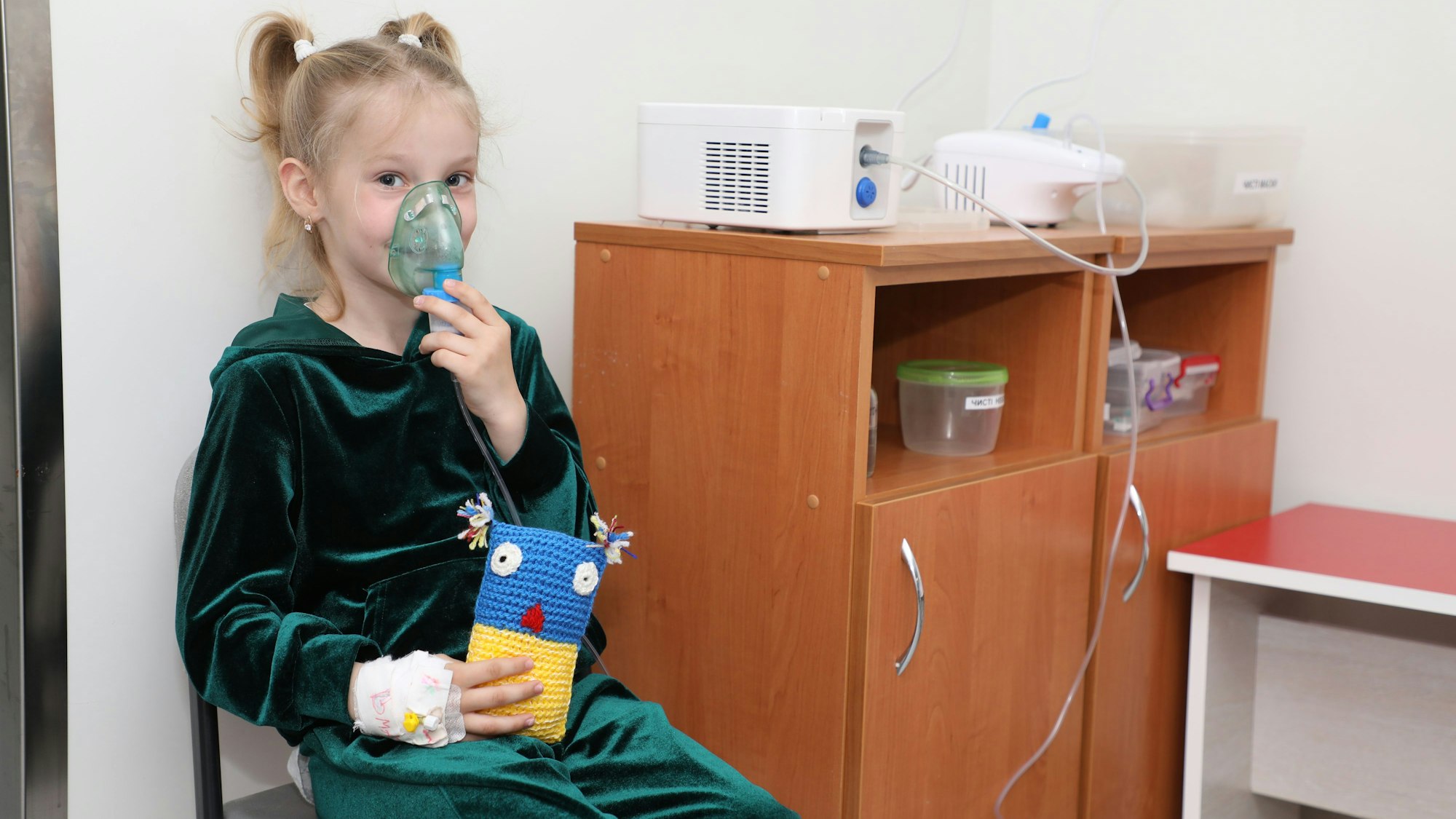 Ein Mädchen mit einem Sauerstoffgerät sitzt auf einem Stuhl und hält ein blau-gelbes Stofftier in der Hand.
