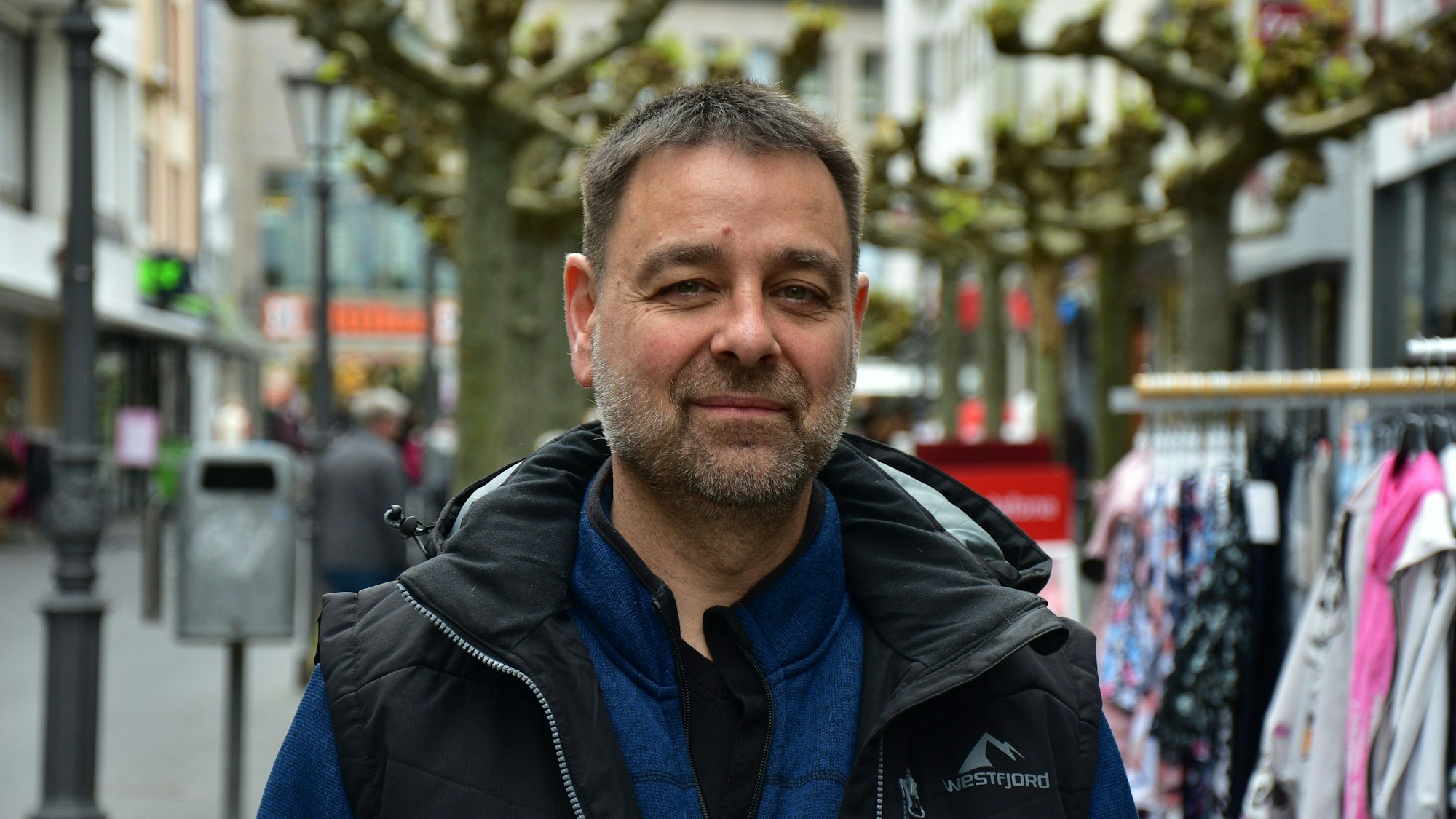 Ein Mann mit Bart in einer Fußgängerzone. Er trägt eine blaue Jacke und darüber eine schwarze Weste. im Hintergrund stehen Platanen und Ständer mit Kleidung vor Geschäften.