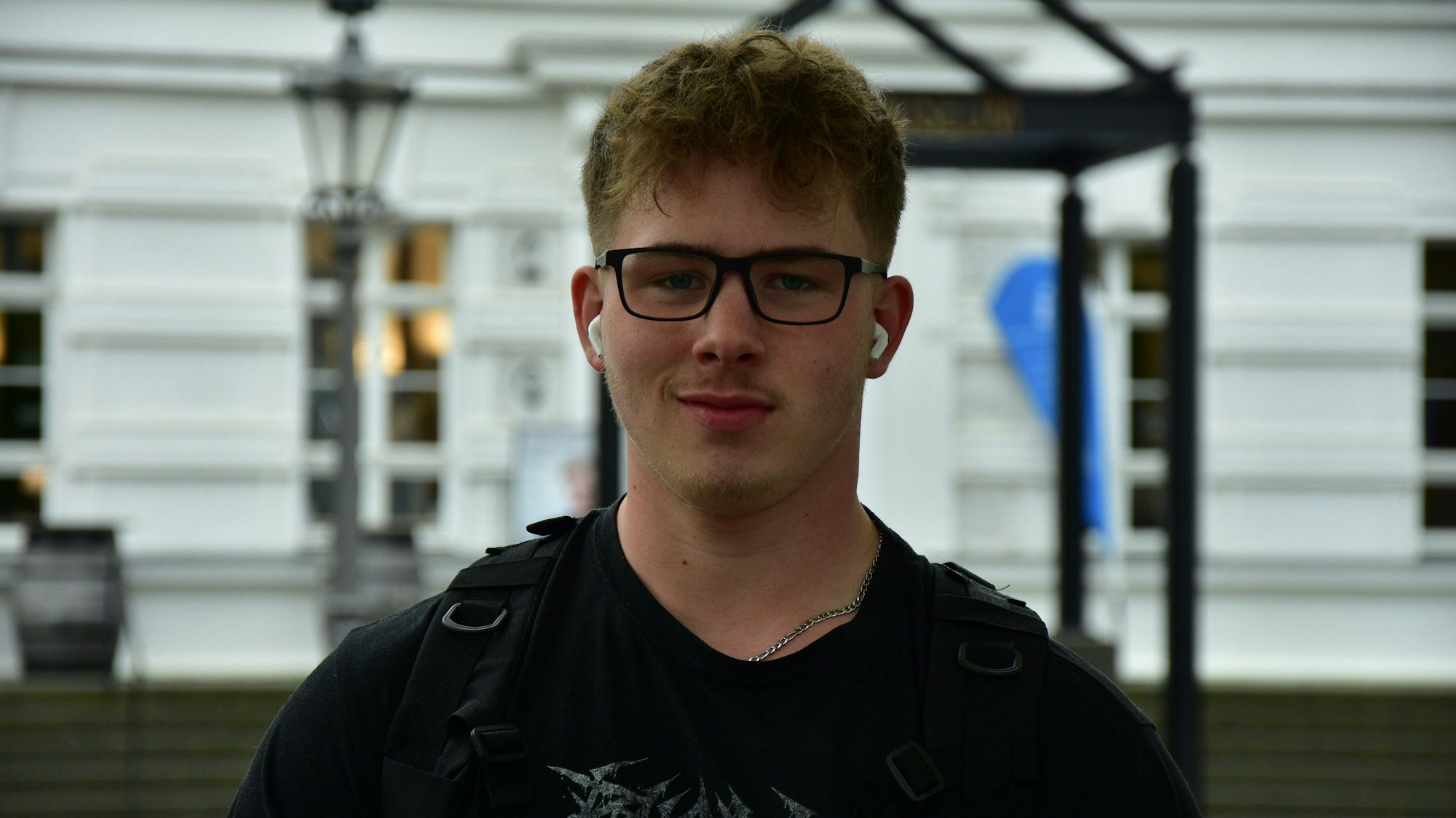 Ein junger Mann mit Brille, schwarzem T-Shirt und schwarzer Jacke vor einem weißen Gebäude. Er hat Kopfhörer in den Ohren.