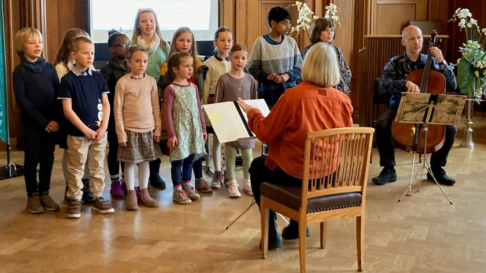 Die Kinder stehen in zwei Reihen hintereinander. Die Musiklehrerin sitzt auf einem Stuhl davor, mit einem Notenständer.
