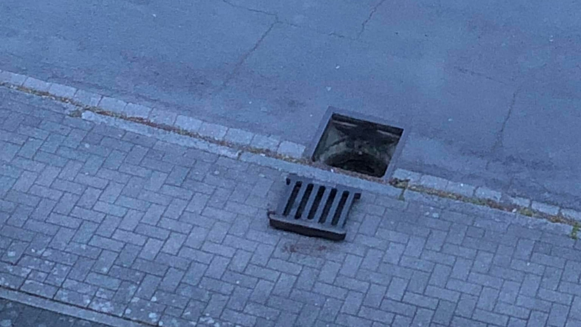 Auf dem Foto ist zu sehen, wie ein Gullydeckel nehmen dem Schacht auf dem Bürgersteig liegt.