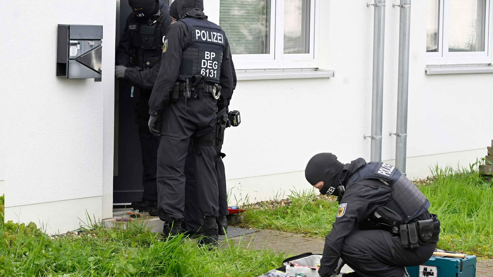 Das Bild zeigt maskierte Polizeibeamte vor einem Haus, das durchsucht wird.