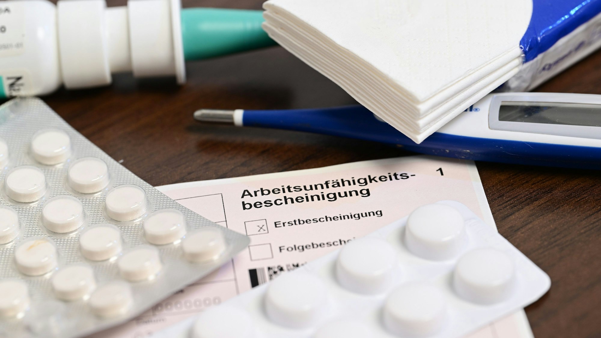 Ein Fieberthermometer, Medikamente und eine ärztliche Arbeitsunfähigkeitsbescheinigung (Krankmeldung) liegen auf einem Nachttisch.