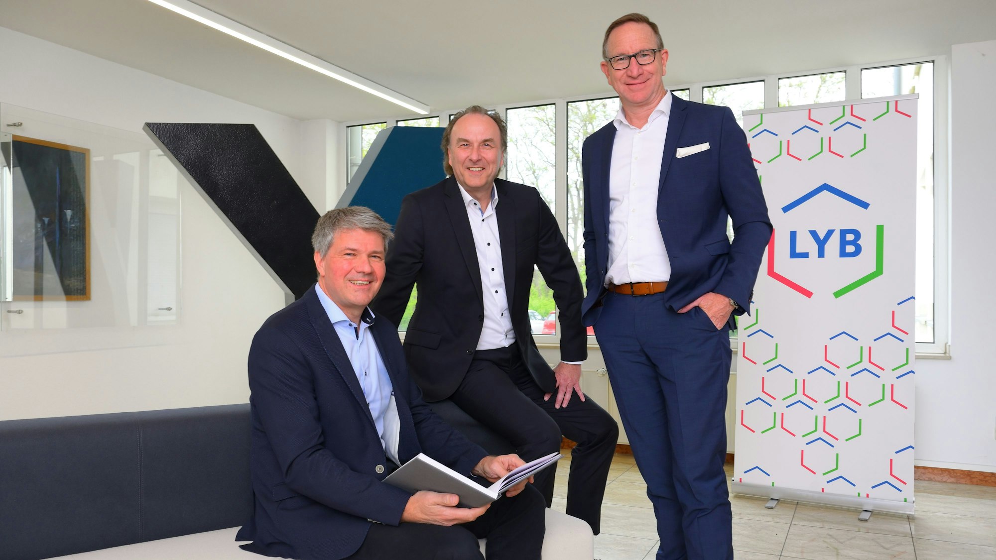 Drei Männer an und auf einem Sofa vor den Firmenlogos der beteiligten Unternehmen.