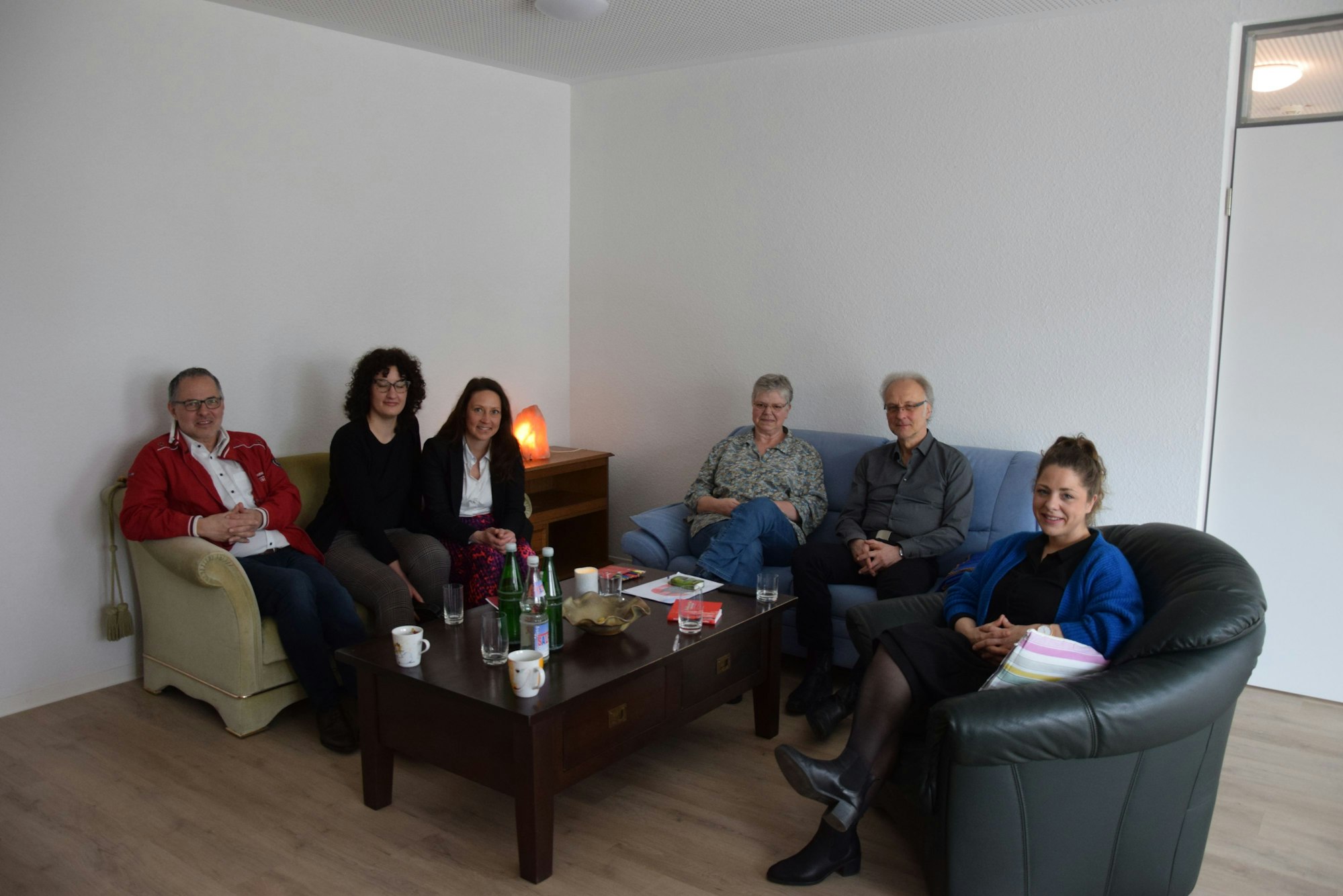 Zu sehen mehrere Menschen, die auf Sofas und einem Sessel sitzen in der Vorderansicht.
