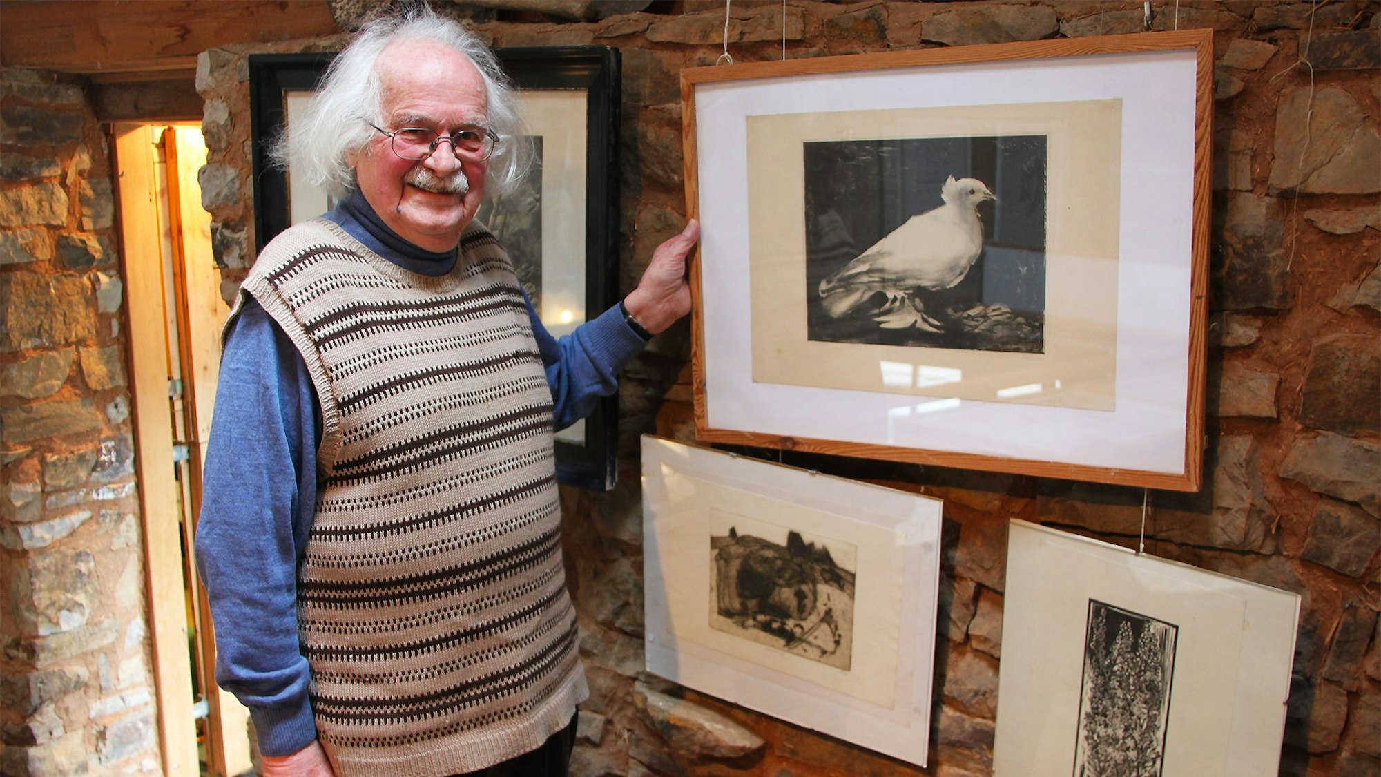 Wolfgang Martens trägt einen Pullunder und steht vor einer Wand, an der mehrere gerahmte Grafiken hängen.