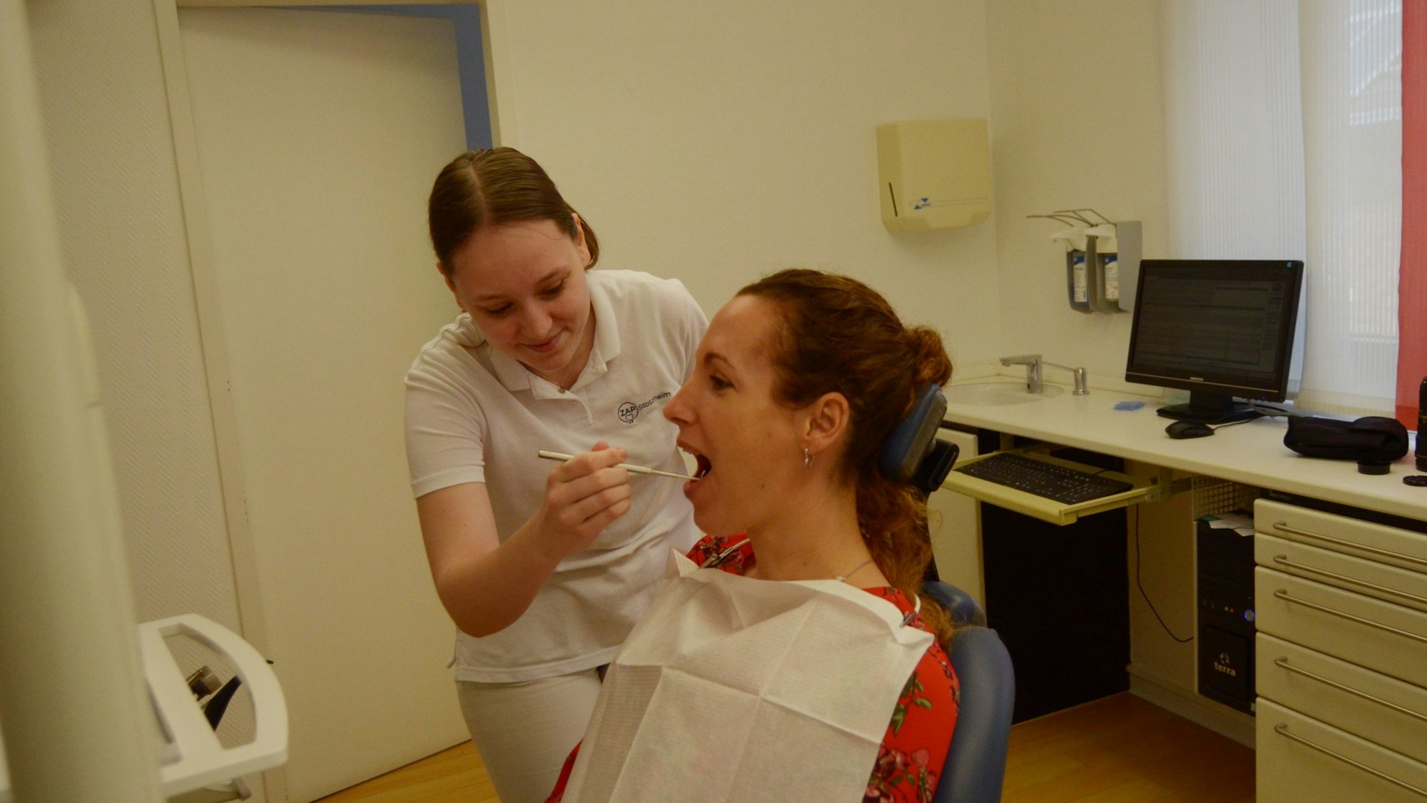 Celine Doerr blickt in der Zahnarztpraxis, in der sie arbeitet, in den Mund einer Patientin, die auf dem Behandlungsstuhl sitzt.