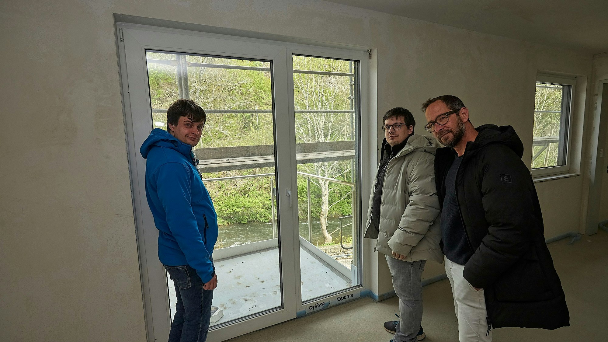 Die drei Männer stehen in einem Raum an der Terrassentür. Unten fließt die Olef vorbei.