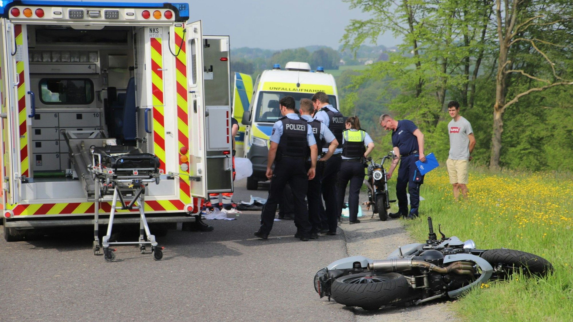 Polizisten stehen an einer Unfallstelle, ein Krankenwagen steht neben ihnen, ein Motorrad liegt auf der Straße.