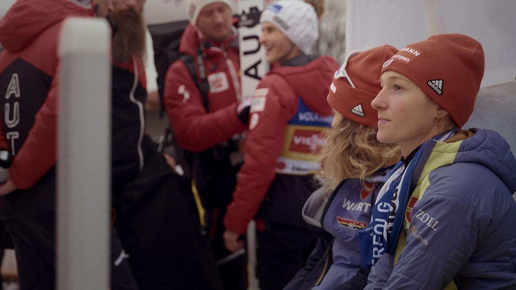 Die Skispringerin sitzt gemeinsam mit anderen auf einer Bank.