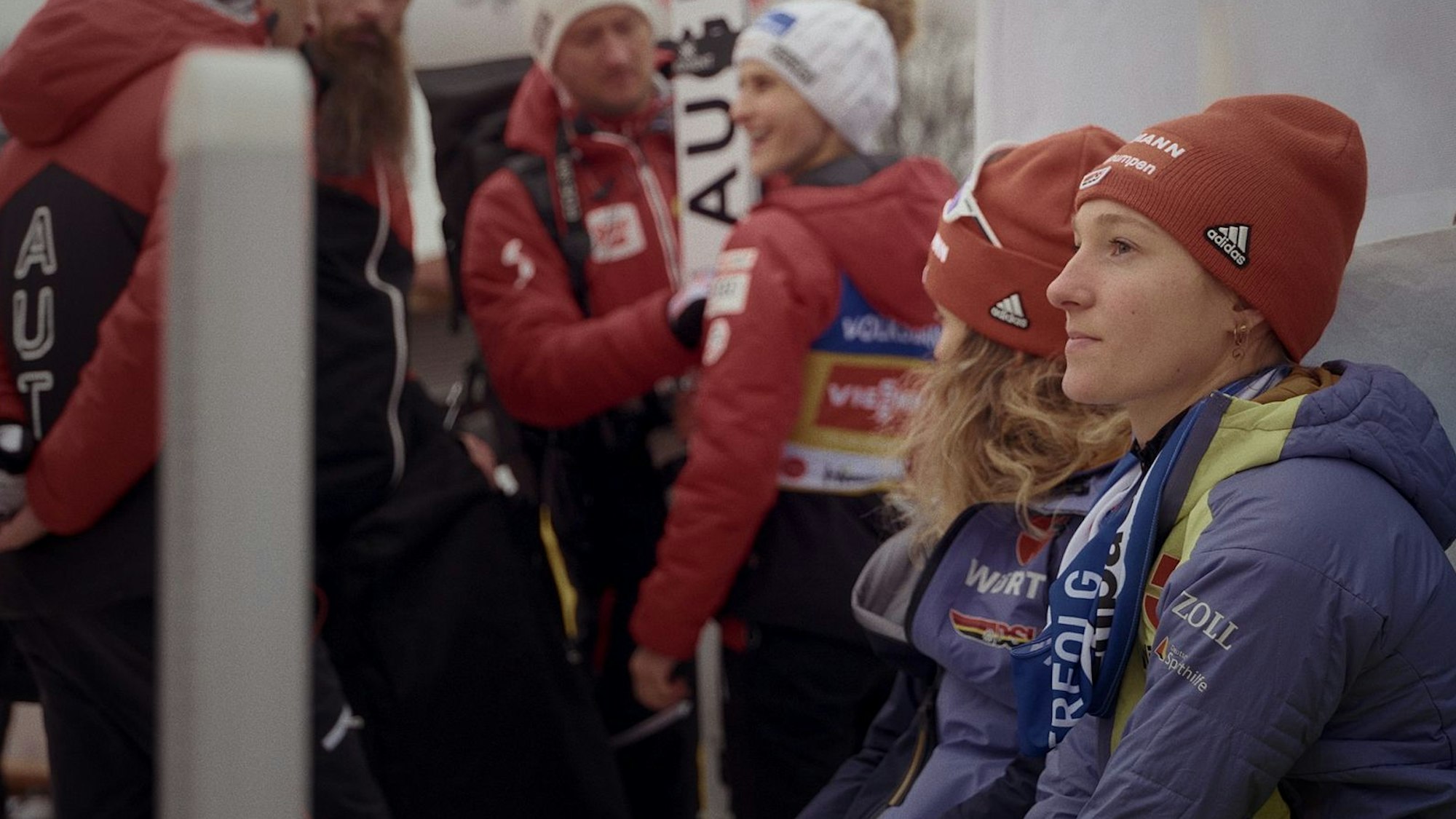 Die Skispringerin sitzt gemeinsam mit anderen auf einer Bank.