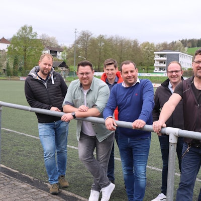 Sechs Männer stehen am Spielfeldrand des Kunstrasenplatzes.