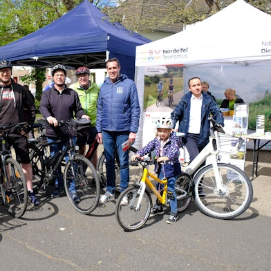 Mehrere Radfahrer, darunter zwei Kinder, stehen mit ihren Fahrrädern in Schleiden vor zwei Zelt-Pavillons.