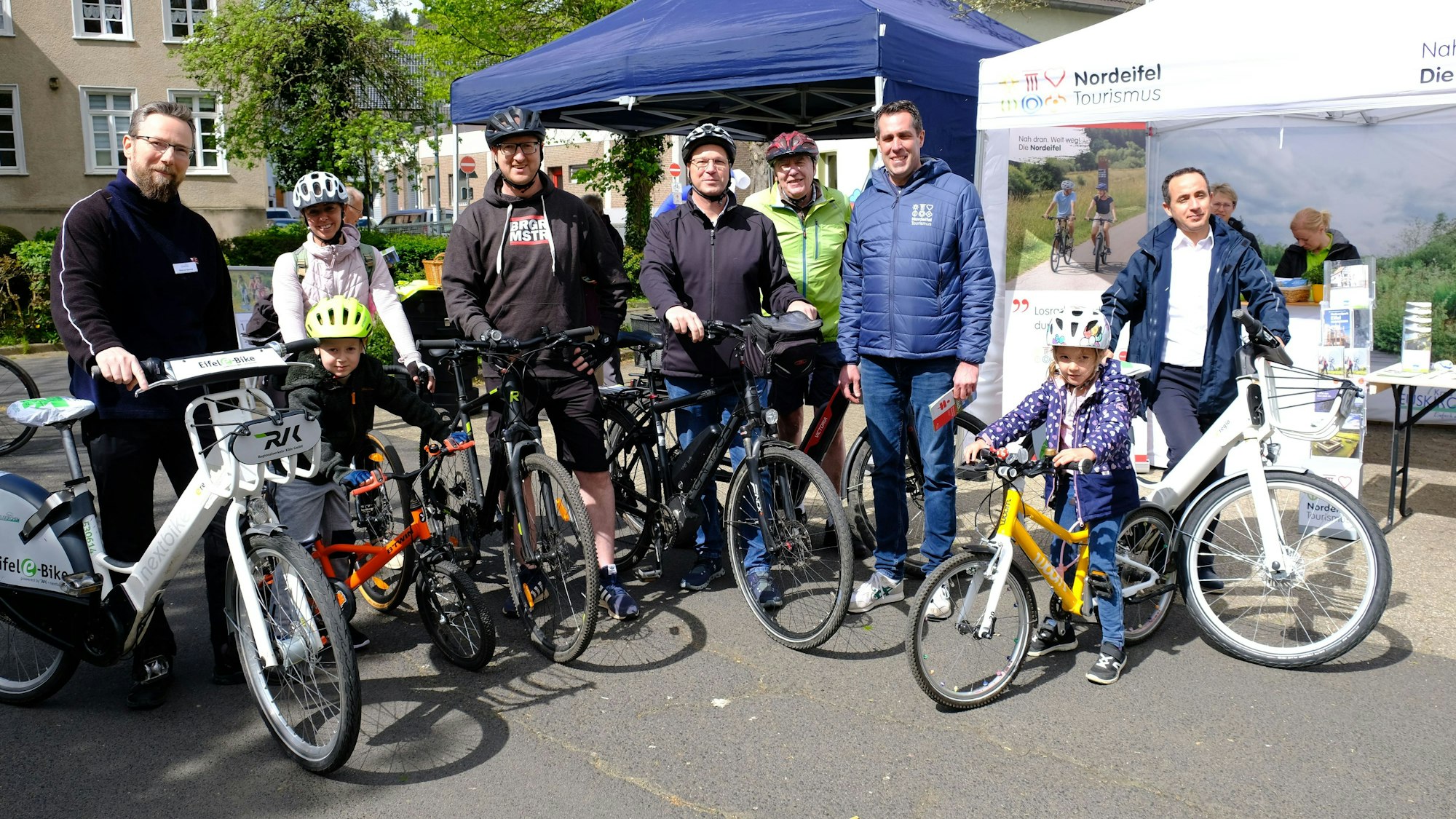 Mehrere Radfahrer, darunter zwei Kinder, stehen mit ihren Fahrrädern in Schleiden vor zwei Zelt-Pavillons.