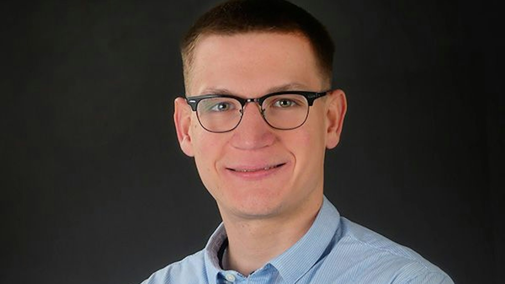 Ein junger Mann mit Brille und blauem Hemd. Er lächelt und schaut geradeaus in die Kamera.