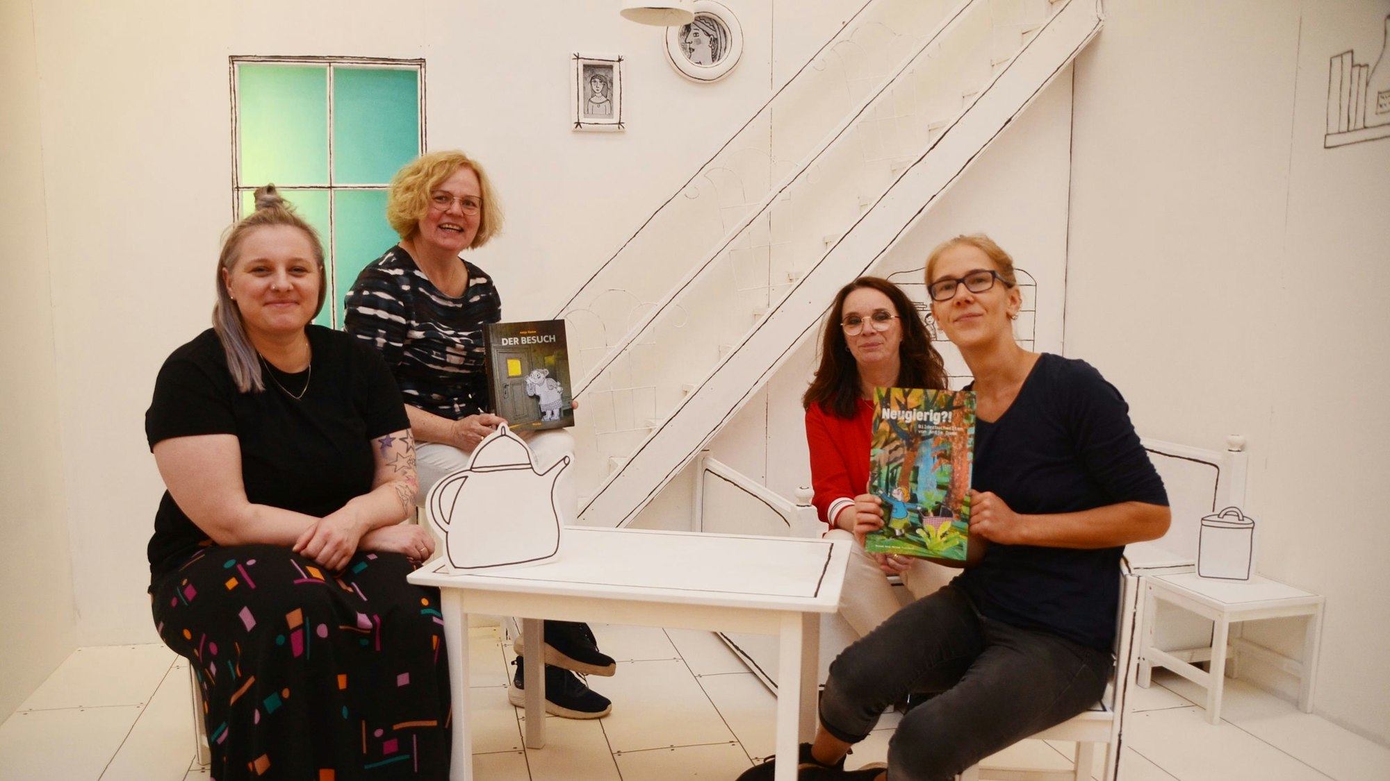 Vier Frauen sitzen auf einer kleinen Bühne, alle Wände und Möbel sind weiß, auch die Treppe oder Bilder an der Wand Die Umrisse sind schwarze Linien.