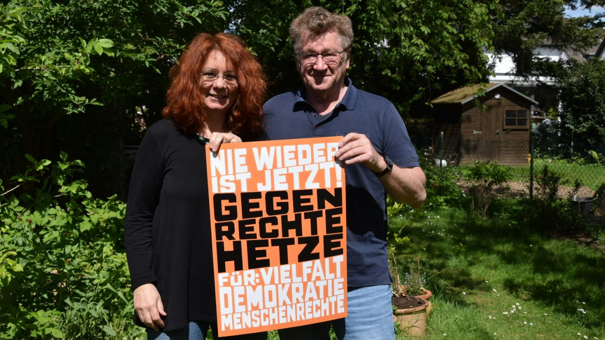Das Plakat gegen Rechts wird in einem grünen Garten hochgehalten von Katrin Lühmann und Willi Vögeli. Darauf ist zu lesen: „Nie wieder ist jetzt. Gegen rechte Hetze. Für: Vielfalt, Demokratie, Menschenrechte“. Foto von Rika Kulschewski