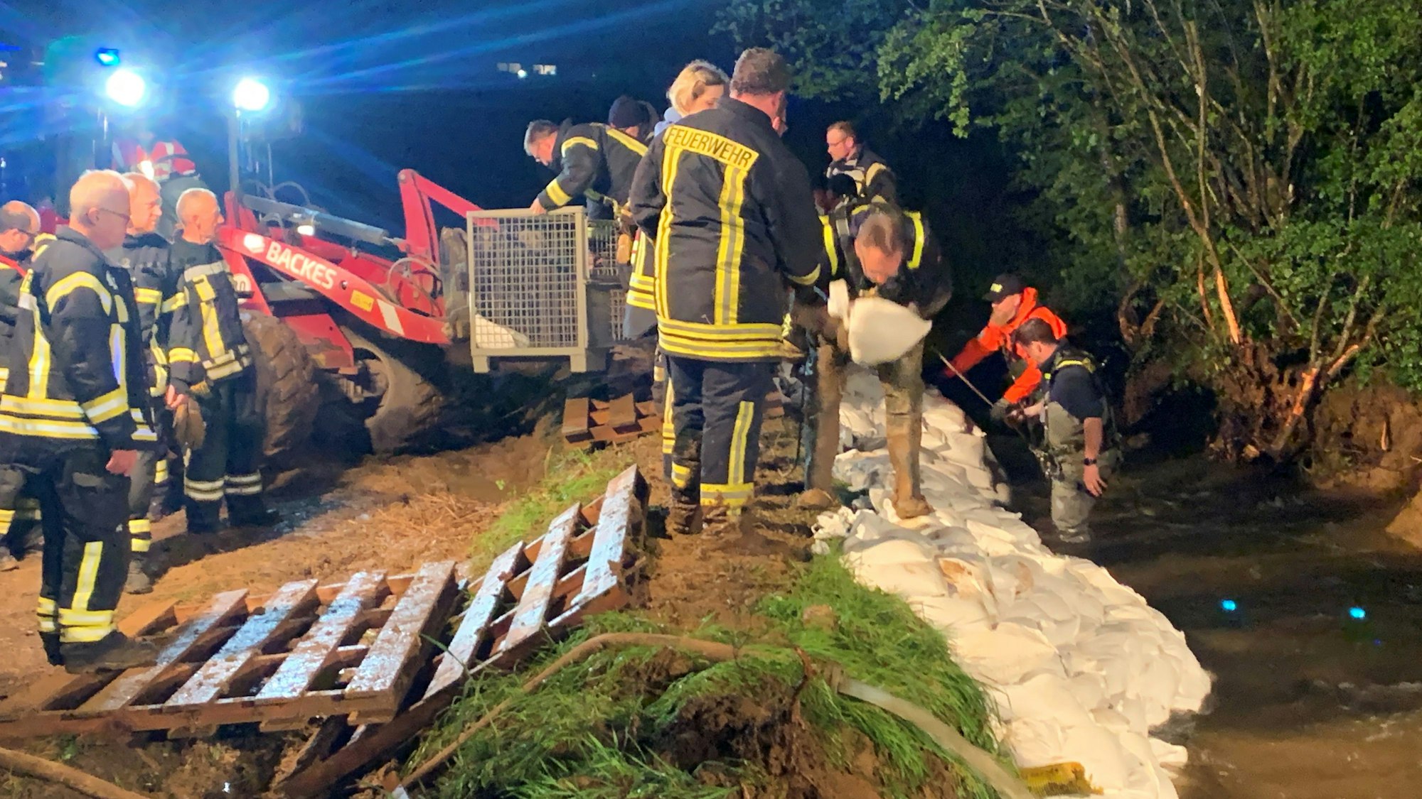 THW-Helfer in Wathosen, an Leinen gesichert, stapeln im reißenden Wasser Sandsäcke, die Feuerwehrleute aus Gitterboxen in einer Kette anreichen.