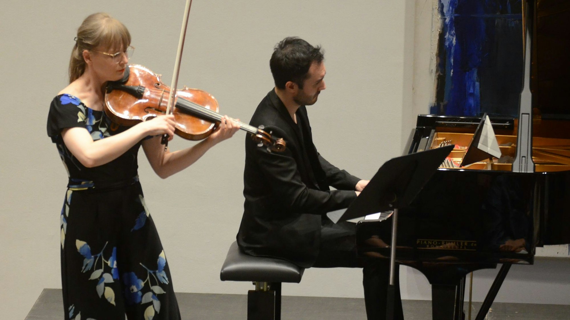 Eine Frau spielt Bratsche auf einem Konzertpodium, ein Mann begleitet sie an einem Flügel.