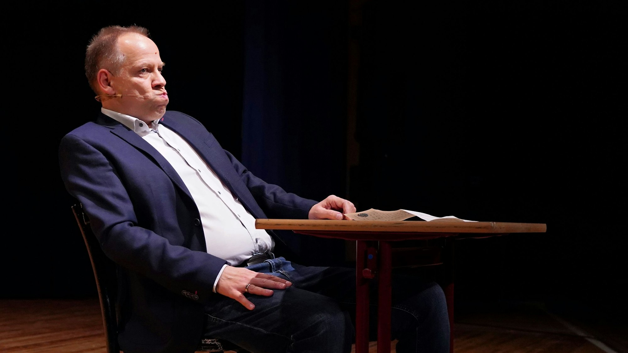 Kabarettist Thomas Schreckenberger sitzt im Stadttheater in Euskirchen an einem Tisch. Er gibt eine gelungene Version von Klaus Kinski zum besten.