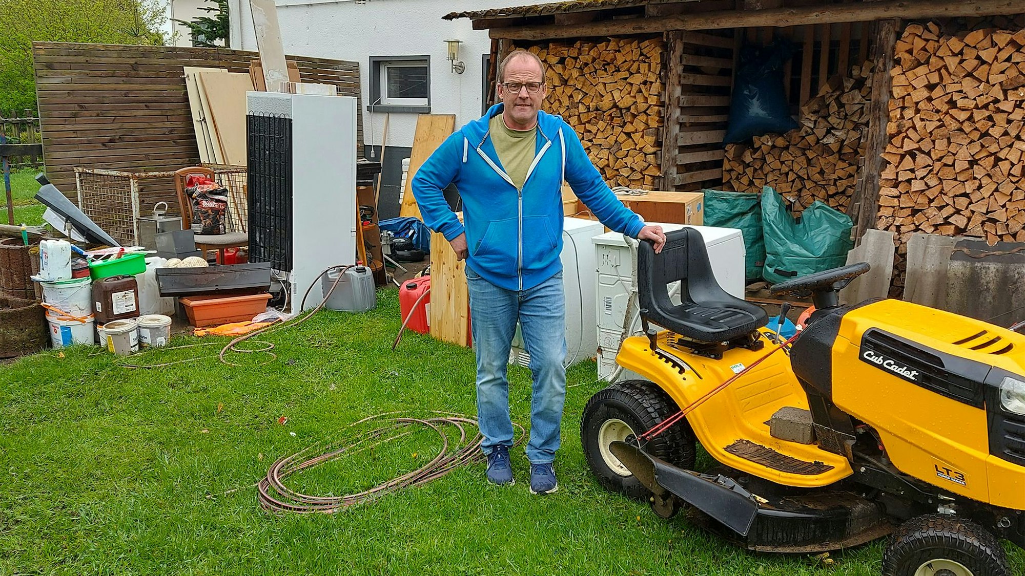 Ein Mann steht in seinem Garten in Dahlem an einem Aufsitzrasenmäher und weiteren Gegenständen, die das Hochwasser zerstört hat.