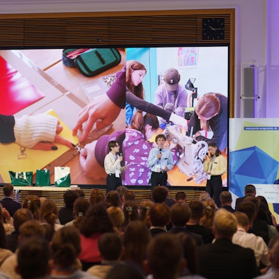 Zu sehen sind eine voll besetzte Veranstaltungsstätte und Moderatoren auf der Bühne.