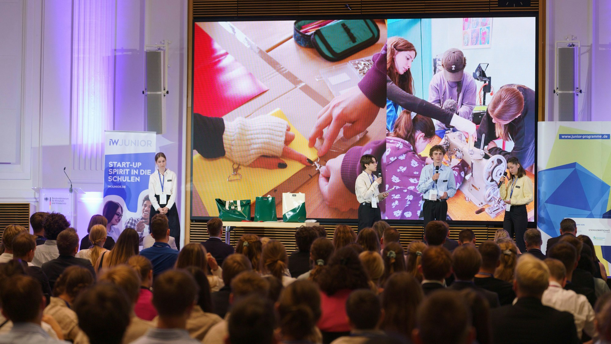 Zu sehen sind eine voll besetzte Veranstaltungsstätte und Moderatoren auf der Bühne.