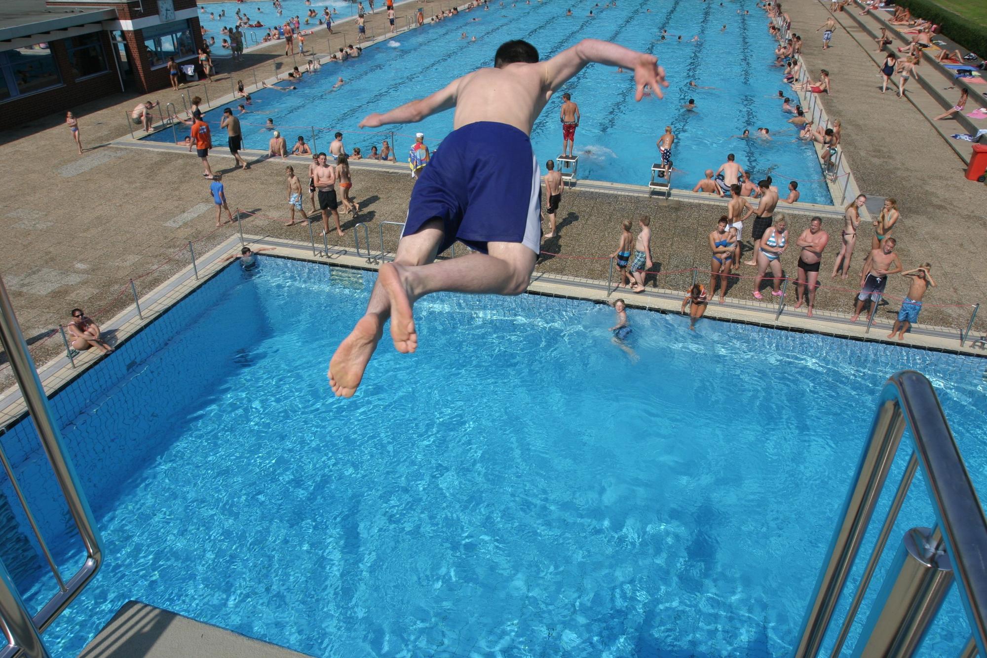 Eine Person springt von einem Turm ins Wasser. Von oben sieht man viele Menschen und zwei Schwimmbecken.