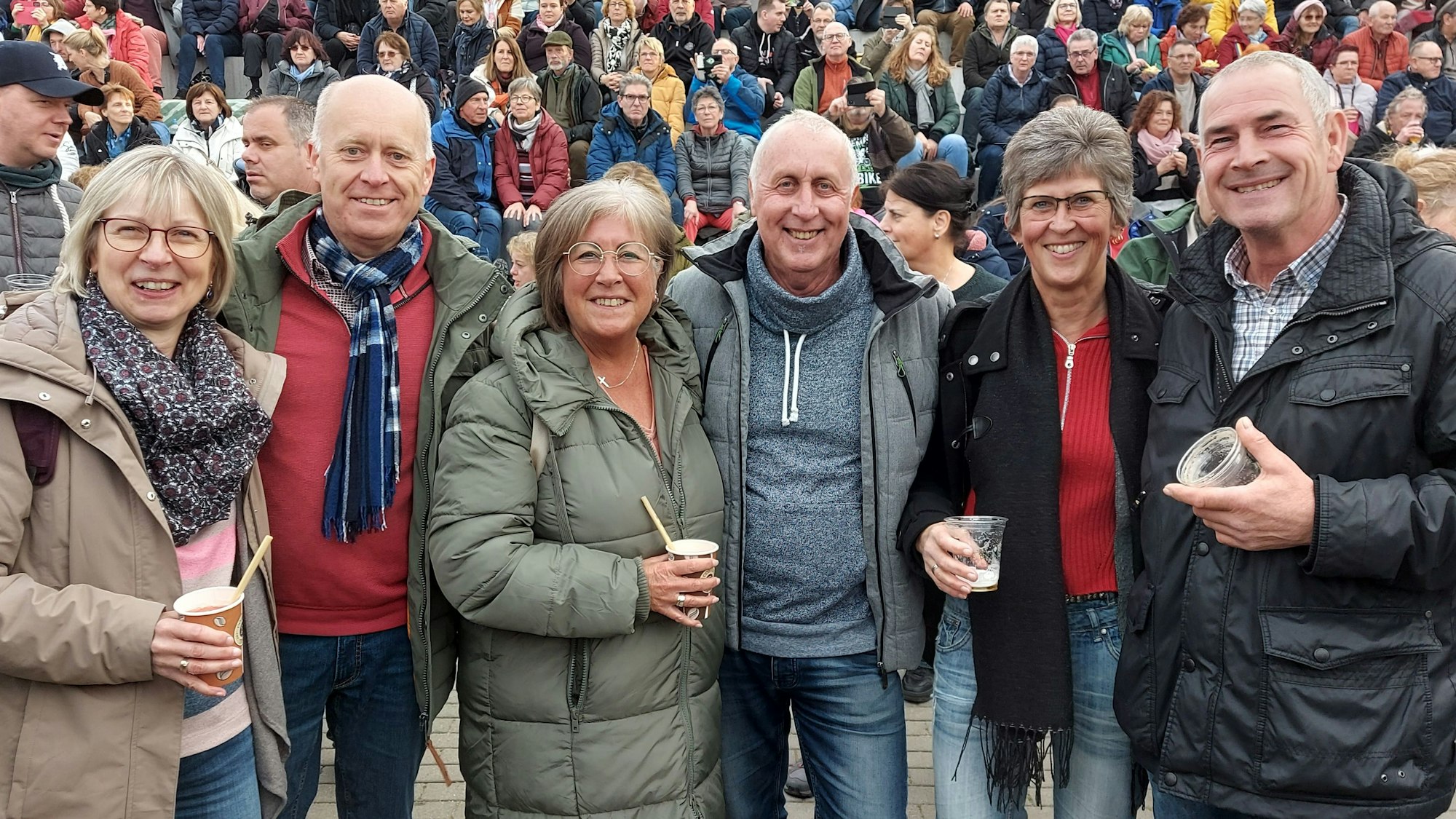 Drei Paare stehen in einer Reihe und lachen in die Kamera, im Hintergrund weitere Zuschauerinnen und Zuschauer.