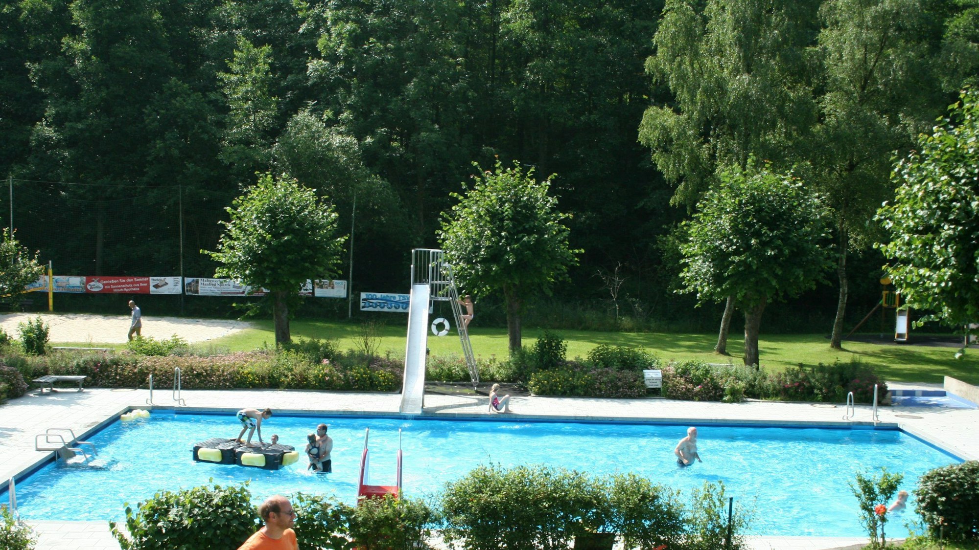 Blick auf ein Schwimmbecken mit Rutsche. Im Hintergrund Bäume und ein Platz mit Sand. Es sind Besucher zu sehen.