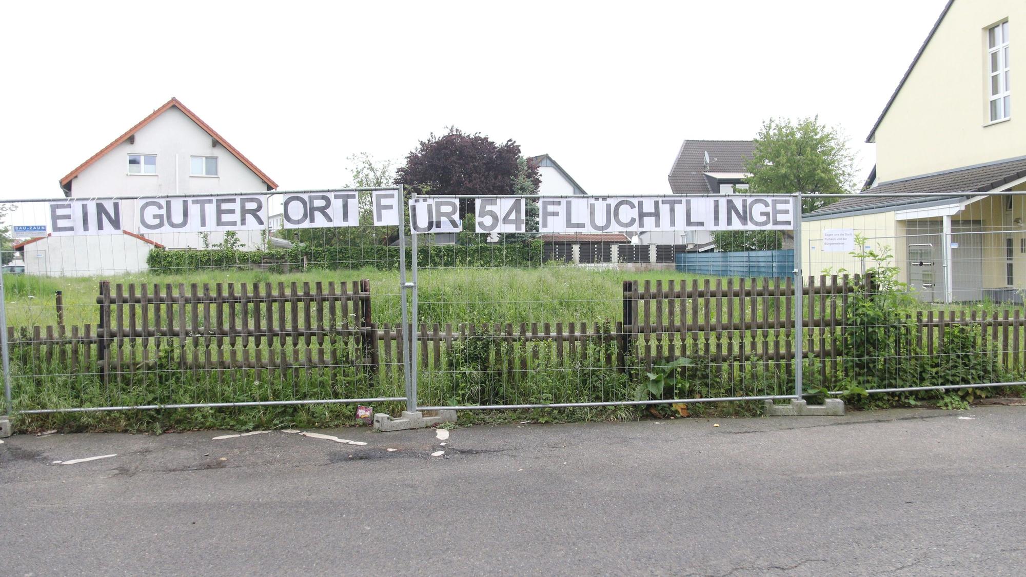 Über einem Zaun hängt ein Transparent mit der Aufschrift „Ein guter Ort für 54 Flüchtlinge“ und daneben ein Zettel mit der Aufschrift „Sagen uns die Stadt Pulheim und der Bürgermeister“.