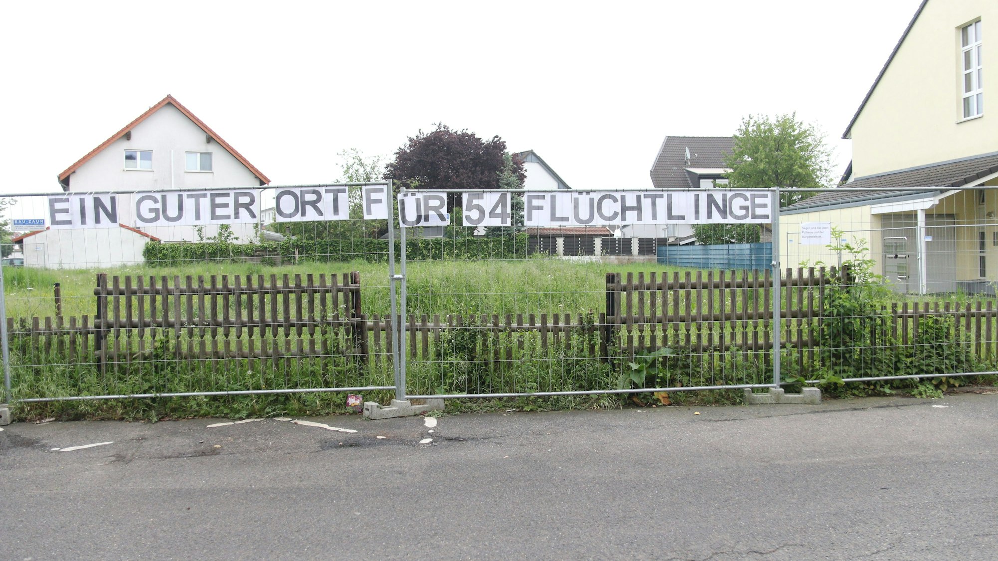 Über einem Zaun hängt ein Transparent mit der Aufschrift „Ein guter Ort für 54 Flüchtlinge“ und daneben ein Zettel mit der Aufschrift „Sagen uns die Stadt Pulheim und der Bürgermeister“.
